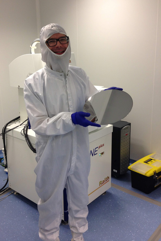 The photo shows Professor Witold Skowroński who is posing in a laboratory with a round polished silicon wafer. The researcher wears a hazmat suit and rubber gloves; there are multiple laboratory devices around him.