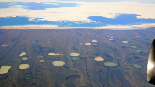 In the foreground, there is a bird’s-eye view of a brown-coloured land dotted with numerous sinkholes. Some of them are filled with azure-coloured water. In the background, there is a fragment of a blue sea, partially covered with white ice floe.