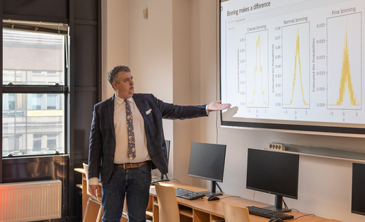 The photo shows Professor Jerzy Baranowski standing in a classroom in front of a projector screen, to which he is pointing with his left hand. The screen shows three yellow graphs.