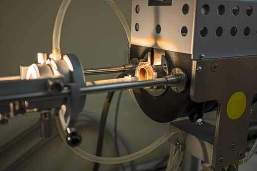 The photo shows a silver high-temperature microscope. In the centre of the photo, there is the sample under investigation, which is illuminated by yellow light that reflects on the microscope as well.