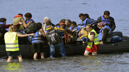 Jednym z powodów wybuchu tzw. arabskiej wiosny były znacznie rosnące ceny żywności Fotografia przedstawia ponton z uchodźcami z Bliskiego Wschodu. Wśród uchodźców są kobiety i mężczyźni. Obok pontonu w płytkiej wodzie stoją też kobieta i mężczyzna w kamizelkach odblaskowych. Mężczyzna trzyma na rękach małe dziecko.