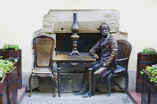 The photo shows the Jan Zeh monument in Lviv. It consists of a table with a paraffin lamp on it and two chairs standing on two opposite sides. One of the chairs is occupied by a man in a long coat who is leaning on the table with his right hand.