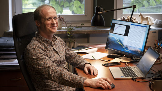 The photo shows the upper part of a young man’s body. He has blonde hair and wears a beige shirt covered in brown print. The man is smiling and sitting in front of a brown desk with computer monitors and a laptop thereon.