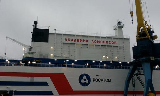 The photo shows a white side of a ship, which has horizontal red and navy blue stripes. There’s an inscription there that says “Akademik Lomonosov” in cyrillic script and an inscription that says “Rosatom” in Latin alphabet.
