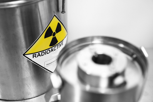 The photo shows cylindrical silver containers for radioactive waste. A black symbol of radioactivity against a yellow background is on the containers.
