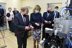 The photo shows a room. On the right side there is a fragment of research equipment. In the middle, there are four people, three of whom (two men and one woman) cut the ribbon with scissors, which spreads in front of the equipment. In the background, on the left, there are four other people standing. There is an inscription on the wall that reads in Polish: Faculty of Foundry Engineering 70 years. All the people in the photo have their mouths and noses covered with protective masks.
