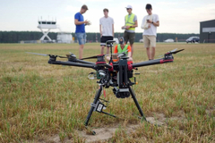Zdjęcie; trawiasta powierzchnia na lotnisku; na pierwszym planie na trawie dron; na drugim planie, za dronem stoi obok siebie czterech młodych mężczyzn, piąty mężczyzna siedzi na trawie. Mężczyźni ubrani są w szorty i t-shirty. Jeden z nich, stojący po prawej stronie, ma na temblaku obandażowana prawą rękę. W tle – budynki lotniska, za nimi gęste pasmo drzew.