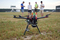 Zdjęcie; trawiasta powierzchnia na lotnisku; na pierwszym planie na trawie dron; na drugim planie, za dronem stoi obok siebie czterech młodych mężczyzn, piąty mężczyzna siedzi na trawie. Mężczyźni ubrani są w szorty i t-shirty. Jeden z nich, stojący po prawej stronie, ma na temblaku obandażowana prawą rękę. W tle – budynki lotniska, za nimi gęste pasmo drzew.