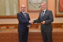 Na zdjęciu dwaj mężczyźni ubrani w garnitury. Podają sobie w uścisku prawą dłoń. W lewej każdy z nich trzyma obwolutę na dokumenty z logo AGH.