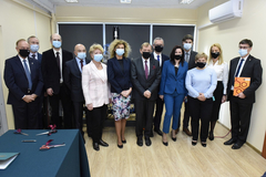 A group photo in a room. There is a row of 13 people standing next to one another. Their mouths and noses are covered with protective masks. In the bottom-left corner, there is a fragment of a table covered with dark blue cloth. There are scissors, a pen, and a paper file with documents on the table.