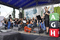 Zdjęcie przedstawia zadaszona scenę. Za sceną widać baner z napisem Dzień AGH. Na scenie ok. 30 osób. Większość stoi, część siedzi na krzesłach i trzyma w dłoniach instrumenty smyczkowe.