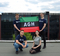 The photo shows members of the Nucleus research group. Two men are holding the AGH flag. When viewed from above, the flag consists of green, black and red stripes. There is a white AGH inscription on the black stripe. At the bottom, the red stripe has the inscription University of Science and Technology in Cracov. Looking from the left, a man and a woman are kneeling under the flag.