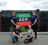 The photo shows members of the Nucleus research group. Two men are holding the AGH flag. When viewed from above, the flag consists of green, black and red stripes. There is a white AGH inscription on the black stripe. At the bottom, the red stripe has the inscription University of Science and Technology in Cracov. Looking from the left, a man and a woman are kneeling under the flag.