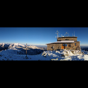 Zdjęcie przedstawia budynek Wysokogórskiego Obserwatorium Meteorologicznego IMiGW na Kasprowym Wierchu na na tle panoramy Tatr. Budynek wykonany jest z szarego kamienia. Ziemia oraz dach budynku pokryte są cienką warstwą śniegu.