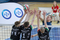 The photo shows a volleyball match. The shot shows three players on one side of the net and one player on the other. All players stick their hands towards the ball that is above the net. The players are represented from the waist up. The three players in the foreground are seen from behind. They have black T-shirts with numbers, surnames, and sponsor names. The AGH UST logo is also visible, as well as the AZS AGH Krakow logo. The player on the opposite side of the net has a navy blue T-shirt. His face is visible. In the background, on the left, there is a logo of PZU – the sponsor of the championships.