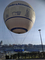 Zdjęcie przedstawia balon widokowy na Bulwarze Wołyńskim w Krakowie. W centrum zdjęcia znajduje się czasza balonu, która jest w kolorze białym. Na balonie znajduje się napis "Dobra Atmosfera" w kolorze granatowym. Pod czaszą zwieszone są liczne liny, który łączą ją z koszem balonu. W tle zdjęcia widać drugi brzeg Wisły oraz stojące przy nim budynki.