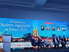 Na zdjęciu widoczna scena, na której w prawej części grupa sześciu osób siedzących na krzesłach. Za nimi wyświetla się slajd konferencyjny.