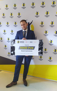 Na zdjęciu młody mężczyzna w garniturze. Stoi na tle białej ściany, na której widnieją logotypy i nazwa konkursu "Studencki Nobel". W dłoniach trzyma planszę z napisem: 5000 PLN nauki techniczne. Na planszy widoczna jest także nazwa konkursu z logotypem.