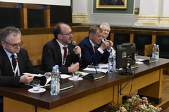 Zdjęcie wykonane w Auli AGH. Widoczne prezydium, przy którym siedzi czterech mężczyzn ubranych w garnitury. Jeden z nich trzyma w ręku mikrofon, inny - broszurę. Na stole: plastikowe butelki z wodą, szklanki, kartki, mikrofony, monitor komputera.