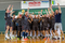 The photo shows the winning male AZS AGH handball team. A group of players stands on the field floor. They wear sports clothes. One of them holds a gold cup. All players cheer for their victory with their hands raised. Their mouths are open, screaming. Their faces look very happy.