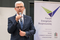 The photo shows a man in a suit who is holding a microphone and speaking to it. Behind him is a board with the Forum's logo.