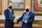 Zdjęcie zrobione w sali konferencyjnej, przed stołem prezydialnym. Na zdjęciu od lewej stoi Prezes Zarządu GroMar, a obok niego Prorektor Rafał Wiśniowski. Obaj mężczyźni są elegancko ubrani w ciemne garnitury, białe koszule i krawaty. Usta i nos mają zakryte maseczkami. W lewych dłoniach trzymają  ciemnozielone teczki z logo AGH z podpisanym porozumieniem, prawe dłonie podają sobie w geście gratulacji. Za nimi, na ścianie wiszą dwie gabloty w których zamieszczone są godło AGH oraz insygnia władzy: berło i topór ceremonialny.