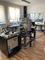 Zdjęcie przedstawia urządzenie stojące w laboratorium. W centralnej części metalowa komora, z której wystają kable i różne rurki. Urządzenie posadowione na metalowych ramach. Po lewej stronie stoi biurko, na którym leży laptop, a pod nim aparatura elektroniczna.