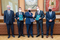 Zdjęcie zrobione w sali konferencyjnej, przed stołem prezydialnym. Przedstawia pięciu mężczyzn stojących obok siebie w rzędzie. Mężczyźni są elegancko ubrani w ciemne garnitury, białe koszule i krawaty. Usta i nos mają zakryte maseczkami. Wzrok kierują do aparatu, w ich oczach widać zadowolenie. Trzech stojących pośrodku trzyma w dłoniach eleganckie teczki z logo AGH z podpisanym porozumieniem, dwaj z nich podają sobie dłonie w geście gratulacji. Za mężczyznami, na ścianie znajdują się trzy gabloty w których zamieszczone są godło AGH oraz insygnia władzy: berło i topór ceremonialny.
