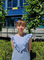 W centrum zdjęcia znajduje się studentka. Studentka ma brązowe włosy. Studentka jest ubrana w niebieską bluzkę i granatowe dżinsy. Studentka stoi na tle budynku i równo przystrzyżonego żywopłotu, którego liście są w kolorze zielonym.