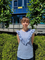 Zdjęcie przedstawia studentkę Dominikę Ciupek. Dziewczyna znajduje się w centrum kadru. Stoi na tle zielonego żywopłotu. Ma blond włosy. Ubrana jest w błękitną bluzkę bez rękawów z falbankami na ramionach.
